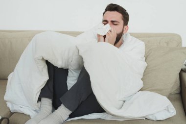 Grip ya da kovid19 'lu genç adam oturma odasındaki kanepede tek başına otururken mendile ya da kağıt mendile öksürüyor.