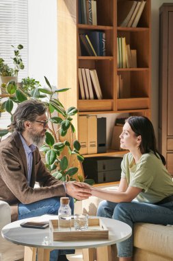 Sakallı kıdemli profesyonel danışman. Genç bir bayanın elini tutuyor. Önündeki koltukta oturuyor.