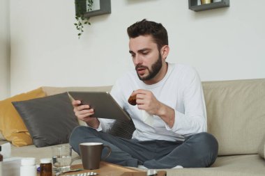 Grip ya da koronavirüs hastası genç bir adam ilaç şişesi tutuyor ve hastalık sırasında evde kalıp online doktora danışıyor.