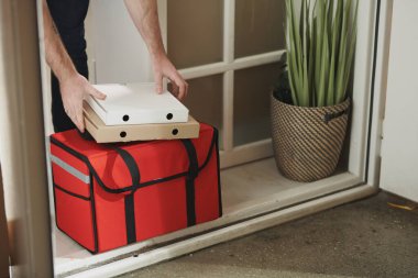Hasta bir gencin elleri iki kutu pizza ve evde otururken yanında yiyecek ürünleri olan büyük kırmızı bir çanta taşıyor.