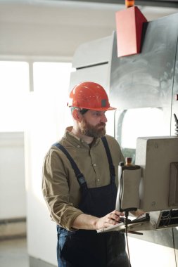 Fabrika atölyesinde çalışırken CNC makine bilgisayarındaki ayarları seçen sıkı şapkalı ve tulumlu sakallı bir operatör.