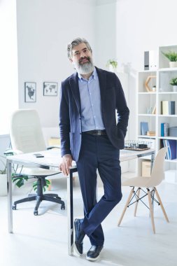 Beyaz deri koltuğun ve ofis raflarının arka planında kameranın önünde duran zarif takım elbiseli yakışıklı bir profesyonel.