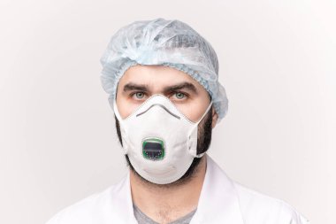 Tıbbi şapkalı genç bir doktor, beyaz yüzgeçli, koruyucu gözlüklü ve solunum cihazlı tecritte kameranın önünde dikilirken sana bakıyor.