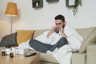 Coronavirus ya da grip olmuş genç bir adam sıcak çay ya da su içerken masanın önündeki koltukta tabletlerle ve haplı şişelerle oturuyordu.