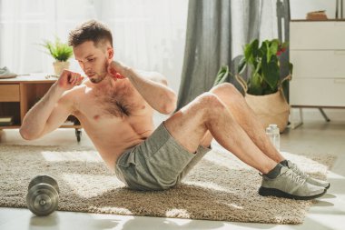 Oturma odasında halıda oturan ve bacaklarını dizlerine kadar bükerek karantina süresi boyunca evde kalan genç üstsüz bir adam.