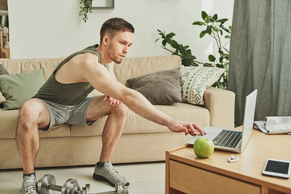 Young sportsman in activewear sitting on couch while stretching arm to laptop to press start button and watch online video of workout