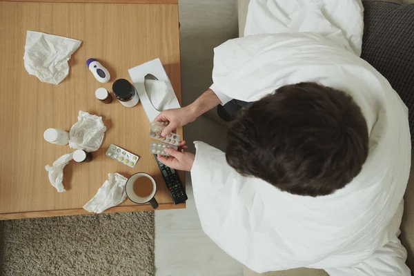 病気の男の概要ソファやテーブルの前のベッドの上に座って毛布に包まれた薬や組織を取るためにどのタブレットを選択する — ストック写真