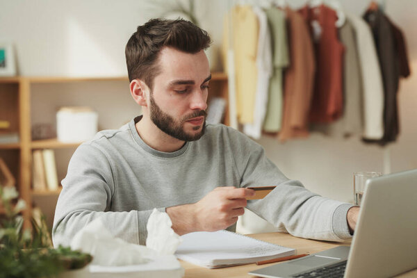 Серьезный молодой бизнесмен смотрит на кредитную карту, сидя перед ноутбуком и собирается заказать лекарства в интернет-аптеке
