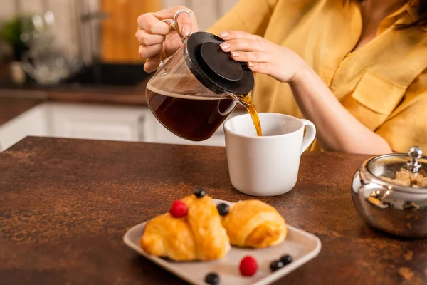 若い女性で黄色のシャツに座ってテーブルとカップに新鮮なハーブティーを注ぎながら 朝食のための新鮮なクロワッサンを持って行く — ストック写真