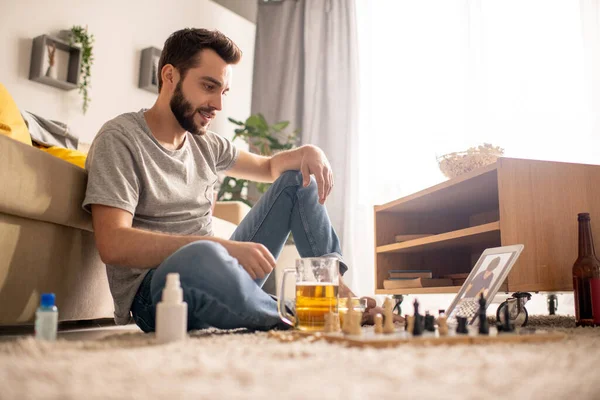在与家人隔离的过程中 积极的 留着胡子的年轻人坐在地板上 手里拿着啤酒杯 与朋友下棋 — 图库照片