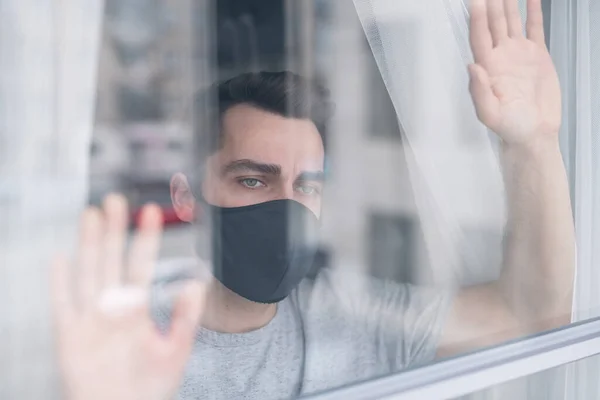 Behind glass view of sad young man in black protective mask holding hands on window and contemplating outdoor scape, he wants to get out of house