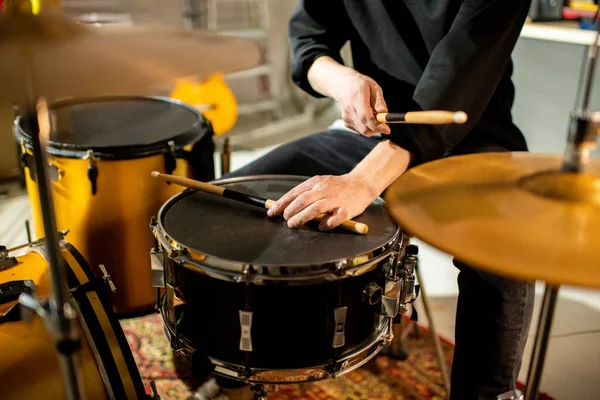 Hand of young musician putting drumstick on black drum while sitting in front of drumset and hitting cymbal during rehearsal in garage
