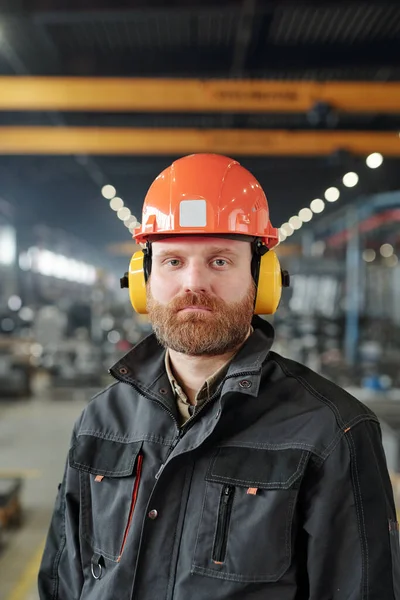穿着工作服 头戴防护帽 严肃的年轻大胡子男子站在工厂车间的画像 — 图库照片