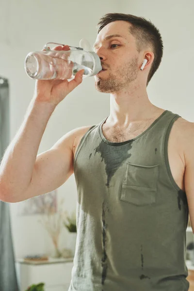 イヤフォンの若い汗びっしょりスポーツマンと隔離期間中に自宅でハードトレーニングした後のボトルから飲料水を着用 — ストック写真