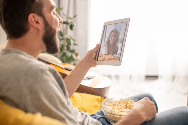 陽気な興奮若いです男食べるポップコーンと笑いながら 隔離中にタブレット上の通信アプリを介してガールフレンドに話をします — ストック写真