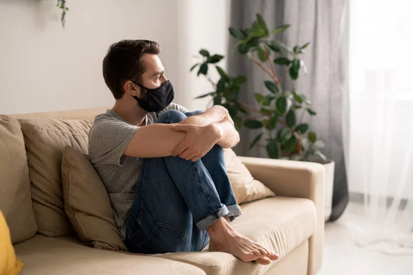 Jovem Nervoso Máscara Facial Abraçando Joelhos Enquanto Sentado Sofá Casa — Fotografia de Stock
