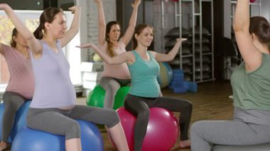 Dört hamile kadın, beden eğitimi dersinde denge toplarının üzerinde otururken kadın antrenörlerinin ardından yoga pozisyonlarını tekrarlıyorlar.