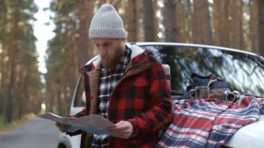 Bereli genç sakallı bir adamın beyaz bir arabanın yanında durup kağıt harita çalışırken orta boy fotoğrafı.