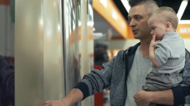 Baba kucağında küçük oğlu, ev aletleri mağazasında koridorda yürüyor ve buzdolabını açıyor.