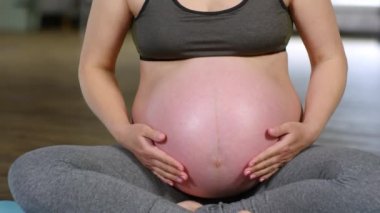 Genç hamile bir kadının yoga minderinde oturup karnını nazikçe ovalarken, kameraya bakıp gülümsediği orta ölçekli bir fotoğraf.