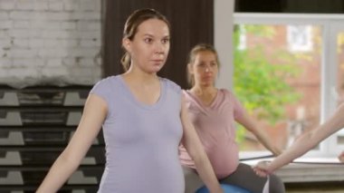 Dengeli topların üzerinde oturan ve spor yaparken bantla omuz daireleri yapan dört aktif hamile kadının orta boy fotoğrafı.