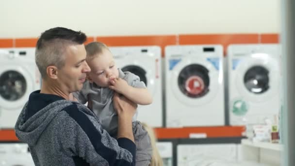 父親は赤ちゃんの息子を腕の上に保持し 2人の娘がクマのおもちゃで遊んでいる間 家電店で洗濯機に彼を置く — ストック動画
