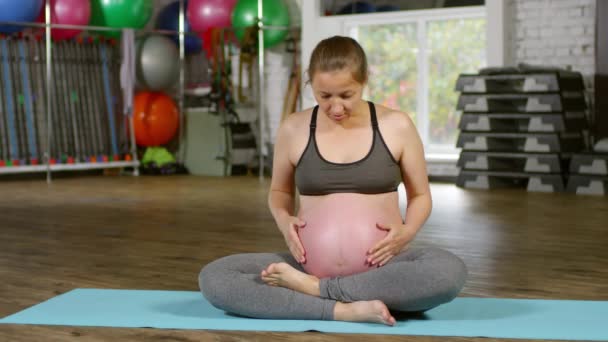 体育館のヨガマットの上に蓮のポーズに座っているスポーツ服の妊婦の肖像画 優しく彼女の腹に触れ カメラを見て — ストック動画
