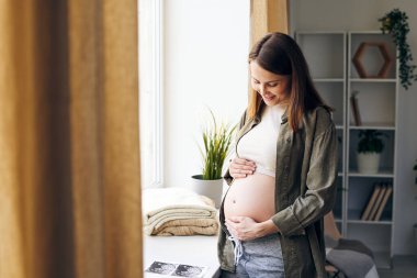 Güzel genç bir kadın hamile karnına dokunuyor ve oturma odasındaki pencereden bebeğini düşünürken gülümseyerek ona bakıyor.