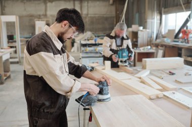 Genç fabrika işçisi öğütücü kullanarak işyeri yüzeyini pürüzsüz hale getiriyor ve daha fazla işlem için ahşap tahtayı hazırlıyor.