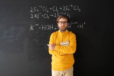 Genç başarılı kimya öğretmeni gündelik kıyafet konusunda çevrimiçi izleyiciler önünde kimyasal formüllerle tahtaya karşı duruyor.