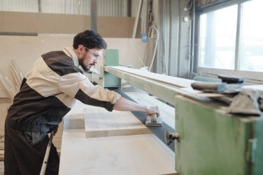 Genç ciddi fabrika işçisi, tezgahın üzerine eğildiği sırada ahşap tahtanın ya da atölyenin yüzeyini pürüzsüz hale getirmek için mekanik el aleti kullanıyor.