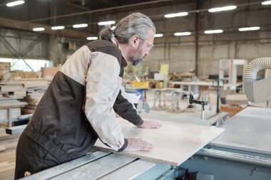 Büyük endüstriyel işleme makinesinin yanında duran ve ahşap tahtanın üzerine eğilerek onu kesen kıdemli işçi.