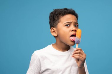 Adorable little boy of African ethnicity licking tasty icecream with taste of juicy orange while looking at you against blue background clipart