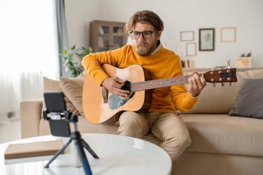 Genç sakallı müzisyen ya da günlük kıyafetlerle müzik öğretmeni oturma odasında kanepede oturuyor ve akıllı telefon kamerasının önünde gitar çalıyor.
