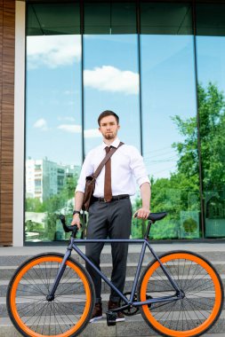 Pantolonlu, gömlekli ve kravatlı genç ve zarif iş adamı modern binanın dış cephesinde merdivenlerde dikilirken bisiklet tutuyor.