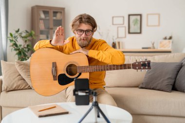 Genç sakallı öğretmen, gitar çalma dersinin başında akıllı telefon kamerasına bakarken çevrimiçi seyircisini selamlıyor.