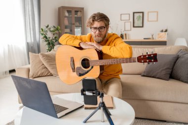 Genç müzik öğretmeni online ders sırasında dinleyicilerine gitar çalarken akıllı telefon kamerasının önünde oturuyor.