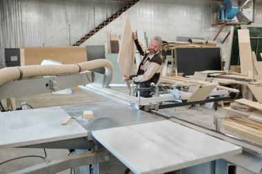 Üniformalı mobilya fabrikasının kıdemli işçisi işlemden önce büyük ahşap tahtaları endüstriyel makineye yerleştiriyor.