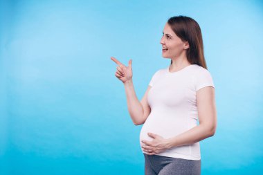 Beyaz tişörtlü neşeli bir kadın hamile karnına dokunuyor ve tecrit edilmiş bir şeye bakarken ileri doğru işaret ediyor.