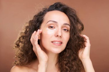 Güzel genç bayan sana bakıyor. Kahverengi arka planda kameranın önündeki göz altı bölgelerine canlandırıcı bantlar koyuyorsun.