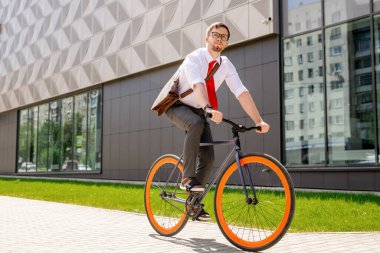 Formalite ve gözlüklü şık bir adam şehir ortamında bisiklet sürerken iş merkezinin dış yüzüne bakıyor.