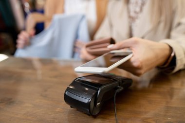Butikten yeni kıyafetler alırken ödeme makinesinin üzerinde akıllı telefon tutan genç bir kadın el.