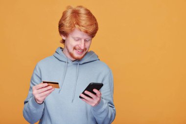 Akıllı telefonlu mutlu genç bir adam çevrimiçi dükkanda malları karıştırıyor ve sipariş verirken kredi kartıyla ödeme yapıyor.