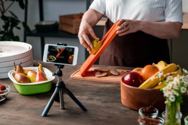 Çağdaş ev hanımı ya da blog yazarı akıllı telefon kamerasının önünde mutfak masasının yanında duruyor ve kuru meyve yapma videosu kaydediyor.