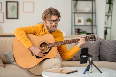 Çevrimiçi gitar çalma dersi sırasında akıllı telefon kamerasına bakan gözlüklü, kot pantolonlu ve kazaklı mutlu genç adam.