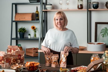 Beyaz tişörtlü mutlu sarışın ev hanımı arkadaşlarına ya da kış için hazırlanmış ev yapımı kurutulmuş meyve paketlerini gösteriyor.