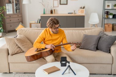 Çağdaş kot pantolonlu genç adam ve sarı kazaklı adam koltukta oturuyor ve evdeki akıllı telefon kamerasının önünde gitar çalıyorlar.