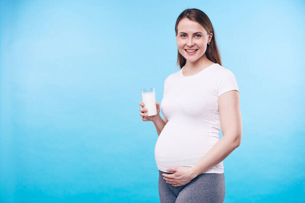 Happy young woman with glass of milk touching her pregnant belly while saying that calcium is necessary for future mother and her baby