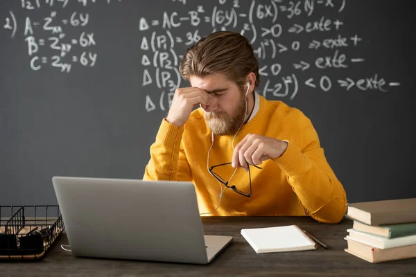 Tired algebra teacher in casualwear touching his face while bending over table in front of laptop during online lesson against blackboard