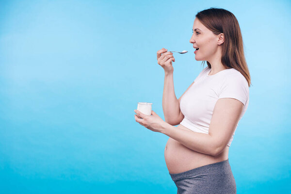 Young active pregnant woman with small plastic cup of dairy product enjoying its taste in isolation with copyspace for your message